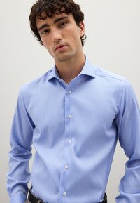 Giovane uomo con capelli ricci che indossa una camicia elegante blu chiaro con bottoni contro uno sfondo semplice e chiaro.