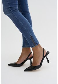 Fekete lakk bőr szandál magassarkú, hegyes orral és 10 cm-es sarkával. Kék farmerral viselve, arany cipzár részletekkel a bokán.