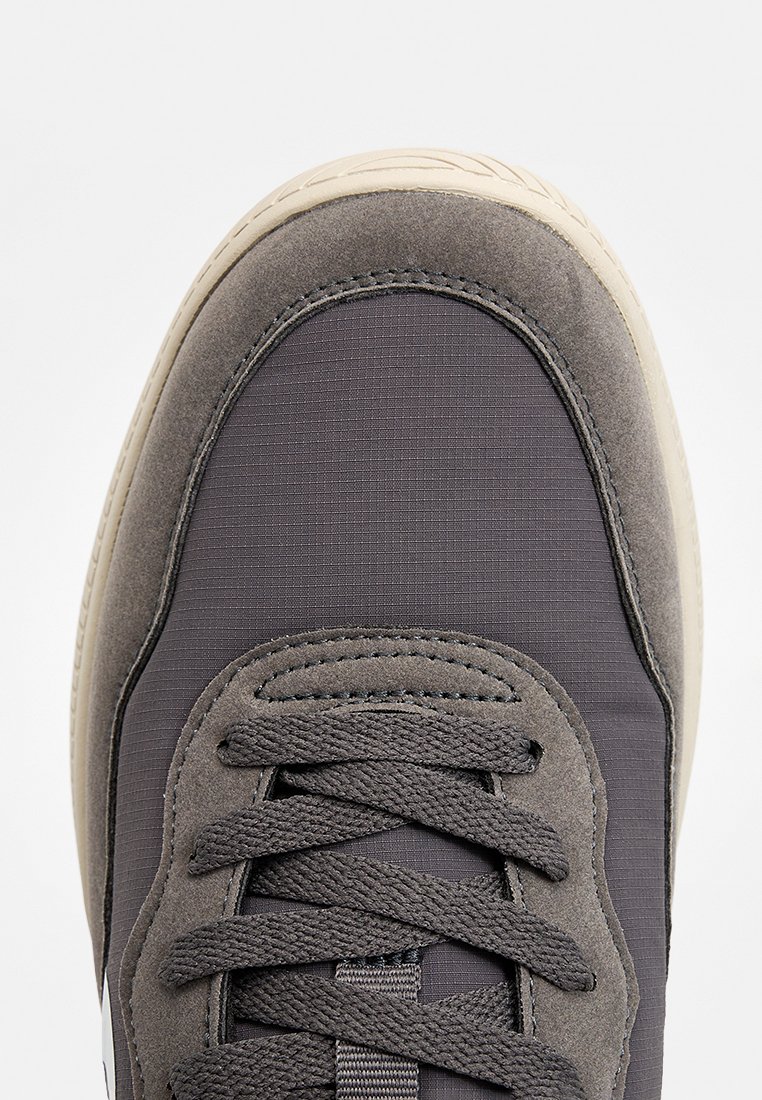 Zapatilla gris con parte superior texturizada que combina ante suave y tejido con patrón. Cordones prominentes y suela de goma. Diseño de punta redondeada.