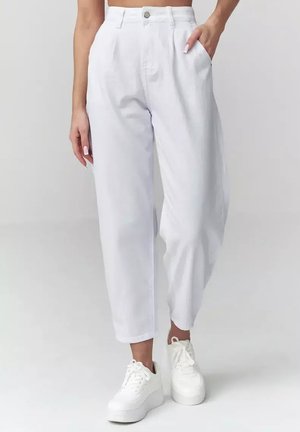 Kobieta w białych, wysokowaschowanych spodniach typu tapered z plisami z przodu i bocznymi kieszeniami, w połączeniu z białymi sneakersami na platformie.