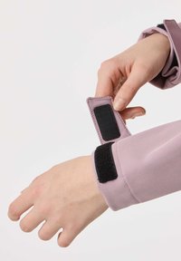 Oseba prilagaja črni ježek trak na manšeti svetlo vijolične jakne na belem ozadju.