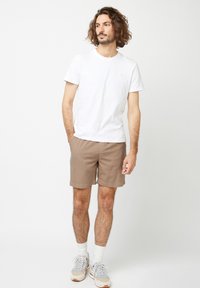 Witte crewneck t-shirt gecombineerd met bruine shorts. Licht materiaal, losse pasvorm. Witte sport sokken en sneakers met lichtblauwe en beige accenten.