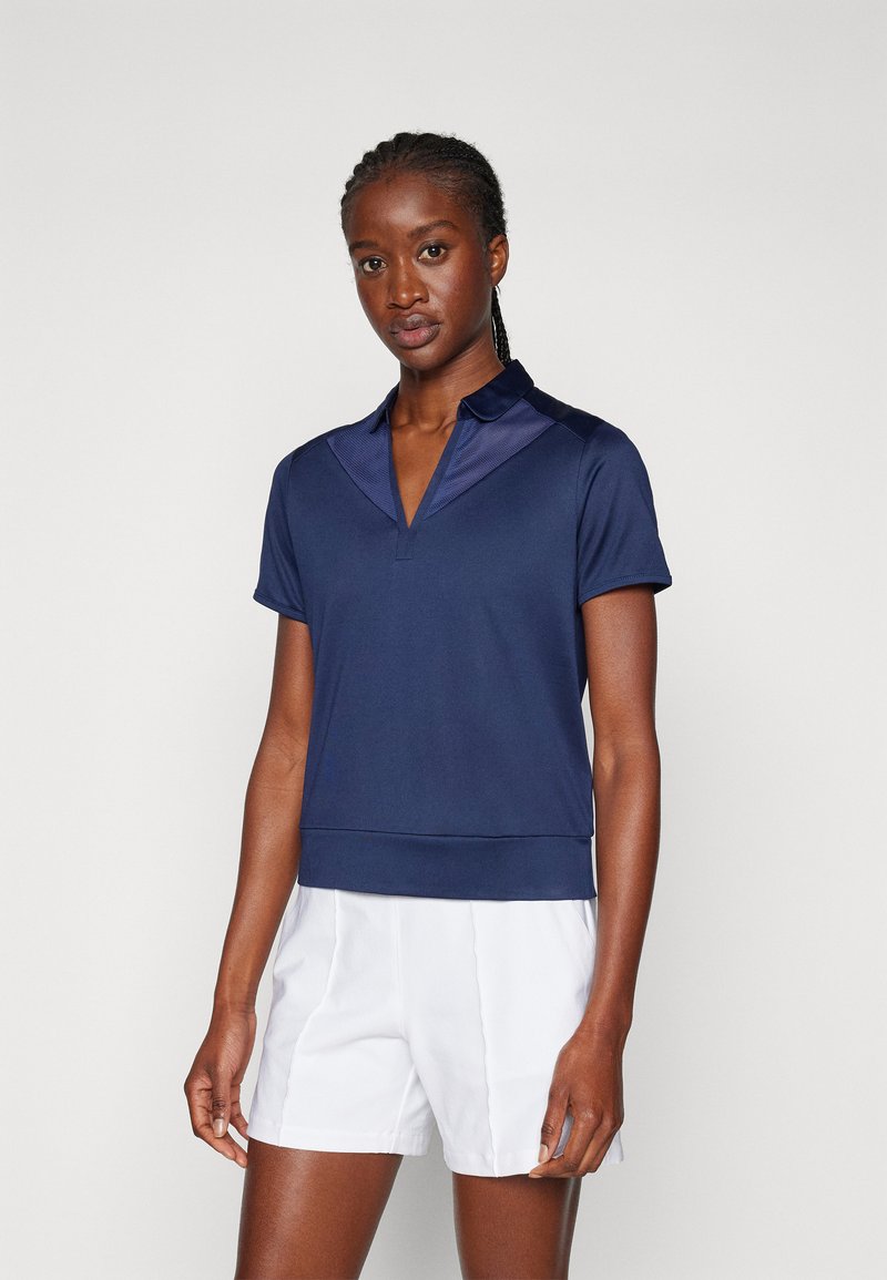 Marineblå kortermet polo med teksturert krage, v-hals og en tettsittende nederkant. Kombinert med hvite shorts, som viser et sporty design.