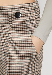 Bavlnené nohavice s houndstooth vzorom v béžovej, čiernej a medenej farbe. Obsahujú bočnú kapsu a čierny gombík v páse.