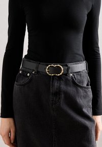 Curea din piele neagră cu o cataramă aurie răsucită, purtată peste un top negru cu mânecă lungă, asociată cu o fustă din denim închis, dotată cu buzunare.