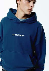 Sininen huppari, joka on valmistettu puuvillasekoitteesta. Siinä on etutasku, nyöritettävä huppu ja "STWDSTUDIO" -logo valkoisena rinnassa.