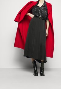 Cappotto rosso di lana con ampi risvolti, blusa a righe bianche e nere, gonna midi plissettata nera e stivaletti neri lucidi con tacco.