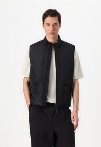Sorte, ermeløs vest laget av lettvevde stoffer, med høy krage, glidelåslommer på sidene og en tettsittende design, båret over en lys topp.