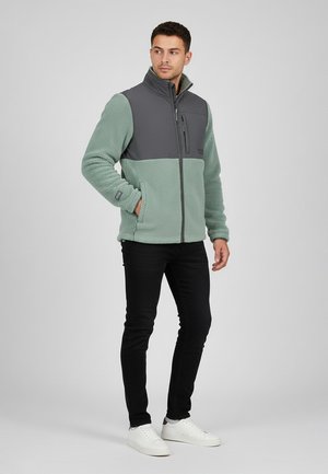 Man standing wearing a green fleece jacket with gray shoulder panels, black pants, and white sneakers, looking to his left.