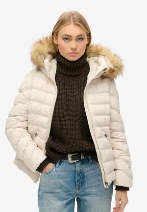 Dikke crème jas met een gevoerde capuchon van bont, gestept ontwerp en een rits aan de voorkant. Gecombineerd met een donkerbruin gebreid sweater en een blauwe jeans.