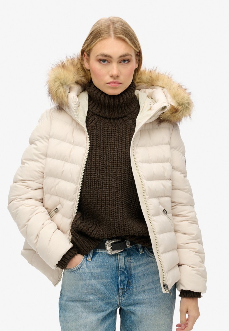 Giacca imbottita di crema con cappuccio foderato in pelliccia, design trapuntato e zip frontale. Indossata sopra un maglione di lana marrone scuro e jeans blu.