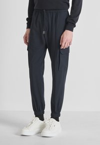 Uomo che indossa pantaloni jogger neri con polsini elastici e tasche, abbinati a sneaker bianche e calze nere, in piedi su un pavimento grigio.