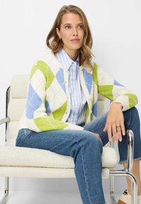 Gebreid vest met een diamantpatroon in groen, blauw en crème. Voorzien van knopen en een geribbelde kraag. Draagt over een lichtblauw gestreept shirt.