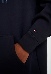 Sudadera azul marino con capucha, que cuenta con un bolsillo frontal y puños acanalados. Pequeño logotipo bordado en rojo, blanco y azul en la manga.