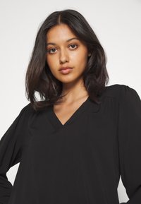 Blusa nera con scollatura a V, maniche lunghe a sbuffo e una consistenza morbida e liscia. Presenta pieghe sottili sulle spalle, per un favorevole adattamento.