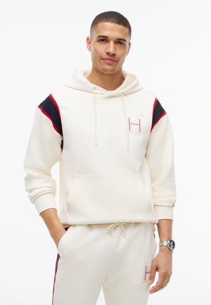 Jeune homme portant un sweat à capuche blanc et un pantalon de survêtement assorti avec des accents rouges et noirs, debout avec une main dans la poche sur fond blanc.