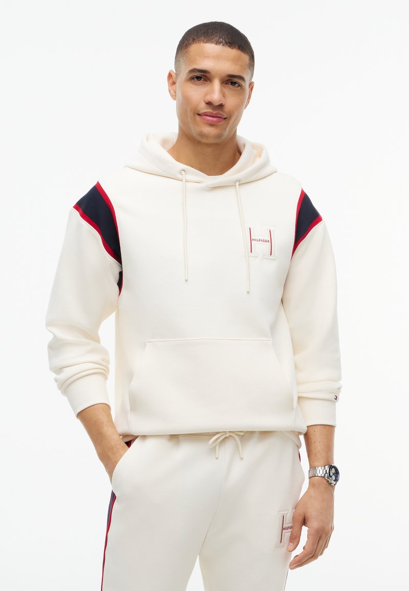 Jeune homme portant un sweat à capuche blanc et un pantalon de survêtement assorti avec des accents rouges et noirs, debout avec une main dans la poche sur fond blanc.