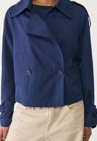 Granatowa, krótka kurtka o podwójnym zapięciu, z ozdobnymi guzikami i subtelnym połyskiem; ma spiczasty kołnierz oraz paski na nadgarstki.