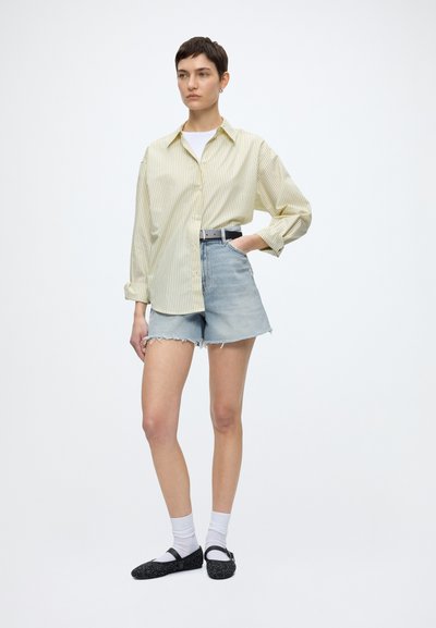 Femme debout avec la main dans la poche, portant une chemise oversize rayée jaune pâle, un short en jean clair, des chaussettes blanches et des chaussures Mary Jane noires.