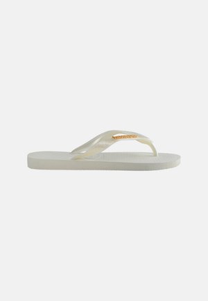 Hvit flip-flop sandal med teksturert såle og gjennomsiktig stropp med en liten gullfarget metall merkeetikett på lys bakgrunn.