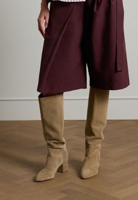 Beige suède kniehoogte laarzen met een blokhak en een zachte textuur, gecombineerd met een bordeauxrode knie-lange rok. Houten vloer op de achtergrond.