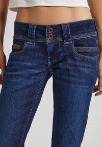 Metade do corpo de uma pessoa vestindo jeans de denim azul escuro com fecho de botão duplo e uma blusa branca cropped, com as mãos relaxadas ao lado.