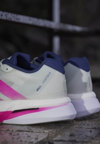 Paire de chaussures de running blanches et roses avec une doublure intérieure bleu marine, étiquetée "ADIZERO BOSTON 13,", posée sur une surface en pierre.