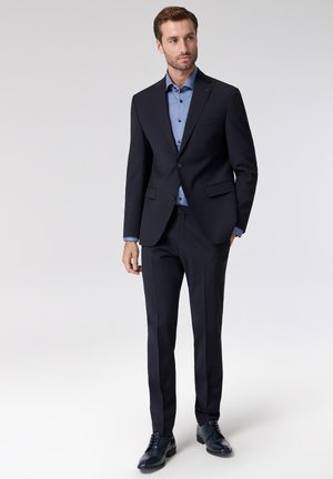 Marineblauer Anzug mit tailliertem Schnitt, passende Hose und hellblauem Hemd. Glatte Textur und dezente Details am Revers. Dunkle Anzugschuhe.