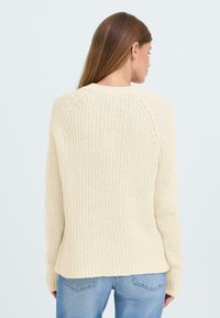 Beige strikket genser med vid hals, ribbestrikkede mansjetter og en lett buet kant. Teksturert stoff med løs passform.