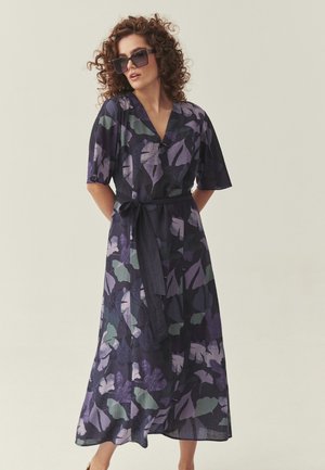 Kobieta o kręconych włosach, nosząca oversize’owe okulary przeciwsłoneczne i ciemną sukienkę w kwiaty z wiązaniem w talii, stojąca na tle gładkiego, jasnego tła.