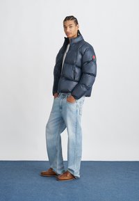 Marineblå puffjakke med høy krage og horisontale sømmer, paret med lyseblå jeans og brune mokasiner, på en blå bakgrunn.