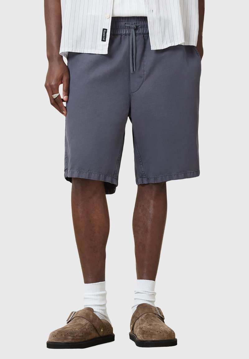 Pantaloni corti grigi realizzati in tessuto liscio, con vita elastica e coulisse, taglio fino al ginocchio e tasche laterali. Indossati con scarpe brown slip-on.