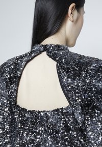 Femme aux longs cheveux foncés portant un top noir et argenté à sequins avec une grande découpe triangulaire dans le haut du dos.