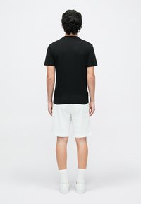 Göndör hajú férfi fekete pólóban, fehér rövidnadrágban, fehér zokniban és fehér tornacipőben, háttal áll egy egyszerű fehér háttér előtt.