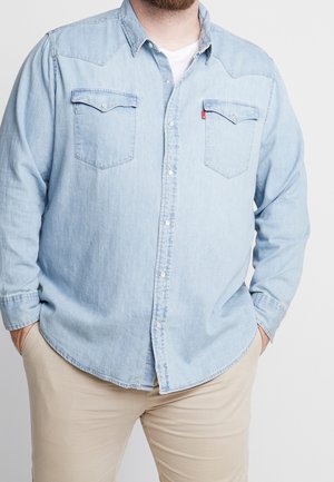 Mann trägt hellblaues Jeanshemd über weißem T-Shirt mit Händen in beigen Hosentaschen, beschnitten am Hals und den Knien.