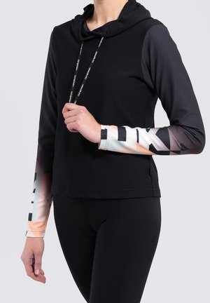 Personne portant un haut noir à capuche à manches longues avec des manches à motif géométrique en blanc, noir et tons pêche, associé à des leggings noirs.