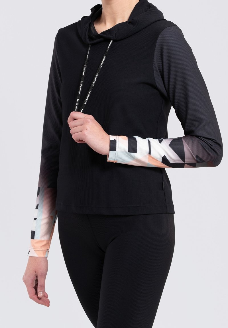 Personne portant un haut noir à capuche à manches longues avec des manches à motif géométrique en blanc, noir et tons pêche, associé à des leggings noirs.