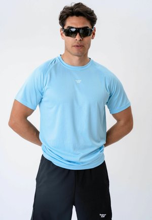 Homme portant des lunettes de sport noires, une chemise à manches courtes bleu clair et un short noir, debout les mains dans le dos.