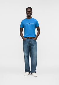 Μπλε βαμβακερό μπλουζάκι με το λογότυπο "TOMMY HILFIGER" σε λευκό, σε συνδυασμό με άνετο μπλε τζιν και λευκά αθλητικά παπούτσια, στέκεται μπροστά σε απλό φόντο.