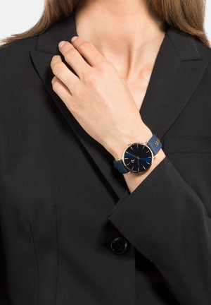 Navyblaue Uhr mit rundem Ziffernblatt, goldenen Akzenten und einem glatten Silikonarmband, am Handgelenk getragen, kombiniert mit einem dunkelblauen Blazer.