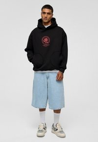 Zwarte hoodie met een rode rozenprint op de voorkant, grote zakken, gecombineerd met lichtblauwe denim shorts. Iets losse pasvorm, casual stijl.