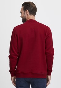 Sarkans džemperis, kas izgatavots no mīksta auduma, ar ribotām kakla un piedurkņu malām, ar brīvu piegriezumu un vienkāršu dizainu, skatoties no aizmugures.