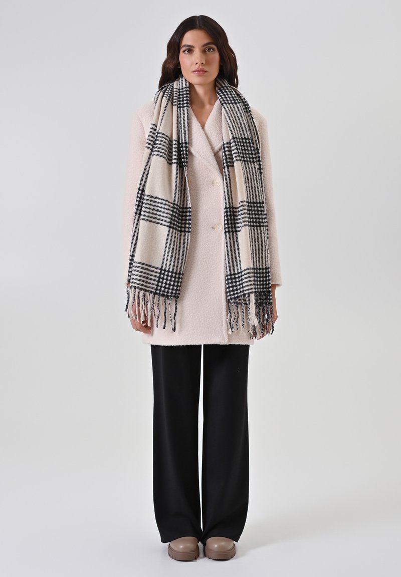 Manteau texturé rose clair avec des boutons doubles, associé à une écharpe à carreaux noir et blanc avec des franges. Pantalon large noir et chaussures marron.