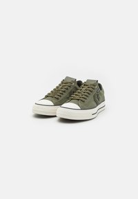 Sneakers in tela verde oliva con puntale e suola in gomma bianca, con dettagli neri e lacci, e una superficie testurizzata.