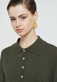 Tamnozeleni pleteni polo majica s šiljastim ovratnikom, s svijetlim, iridescentnim dugmadi i teksturiranim materijalom. Zlatne okrugle naušnice.