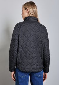 Schwarze gesteppte Jacke mit Stehkragen, versehen mit einem Rautenmuster und langen Ärmeln, kombiniert mit blauen Jeans.