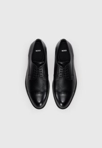 Un paio di scarpe da uomo eleganti in pelle nera lucida con lacci, con il logo "BOSS" all'interno, posizionate su uno sfondo grigio chiaro uniforme.