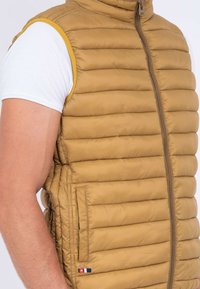 Gilet sans manches en khaki avec texture matelassée, poches latérales et fermeture éclair. Accentué d'une bordure jaune uni le long des emmanchures.