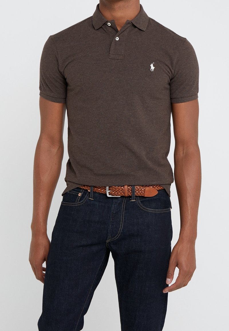 Brun polo med krage, korta ärmar och vitt logo. Parat med mörk denimjeans och ett flätat brunt bälte.