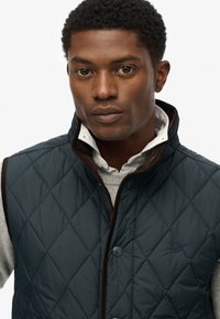 Marineblå quiltet vest med høj krave, knaplukning og kontrasterende brun kant, båret over en lys grå sweater og en hvid skjorte.